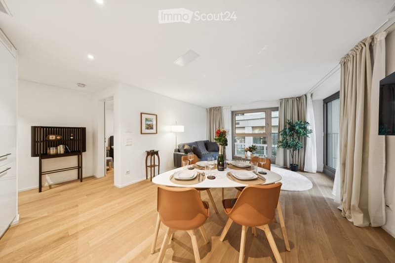 Modern, well-lit living and dining area with wooden flooring, white walls, and large windows. Features a sofa, dining table with chairs, a small cabinet, a table lamp, and decorative plants.
