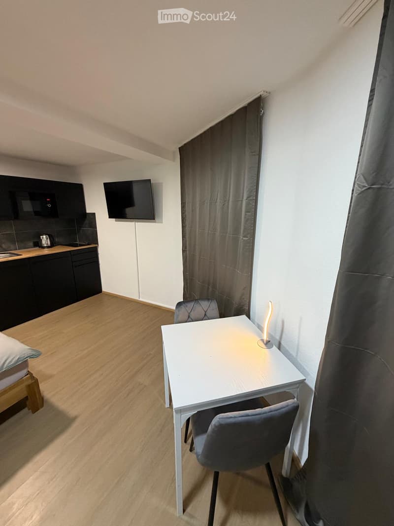 A compact studio apartment with a bed, a small dining table with two chairs, a television, and a kitchenette area with cabinets and a microwave.