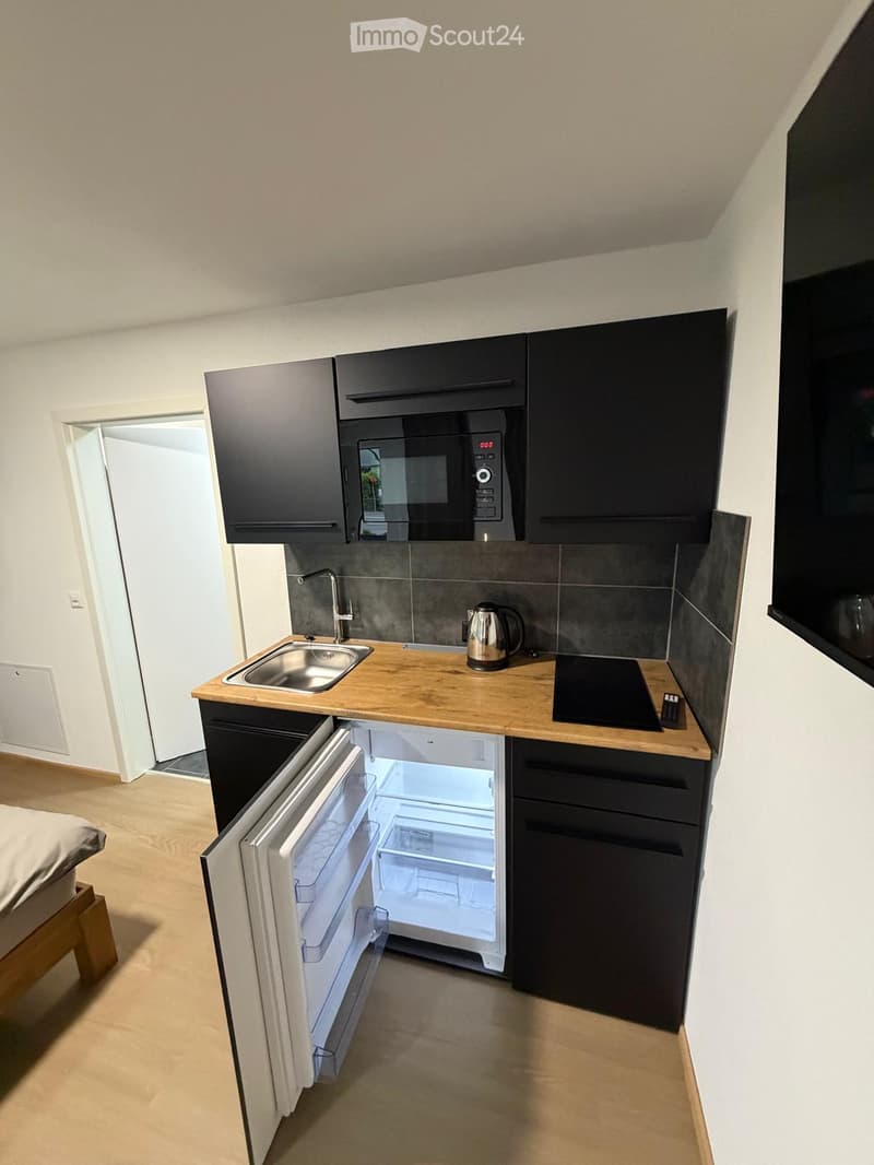 Compact kitchen area with black cabinets, wooden countertop, stainless steel sink, microwave, electric kettle, and an open refrigerator. Adjacent to a bedroom with a wooden bed frame.