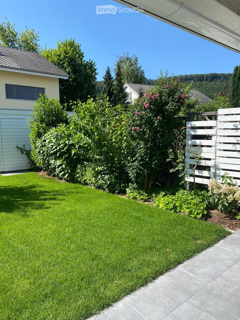 Well-maintained garden with a neatly trimmed lawn, various flowering shrubs, and a white wooden fence. The area is bordered by a concrete pathway and includes a small section of a house with a garage.