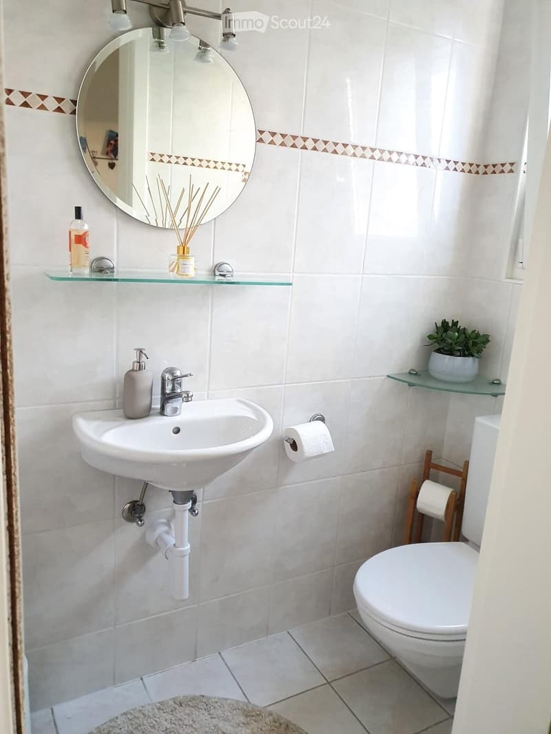 A modern bathroom with white tiles, a round mirror, a sink with a modern faucet, a toilet, and glass shelves holding a plant and toiletries.