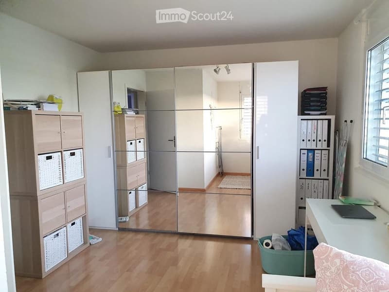 A well-lit room with wooden flooring, a large mirrored wardrobe, a storage unit with baskets, a desk with a laptop, and a small chair.