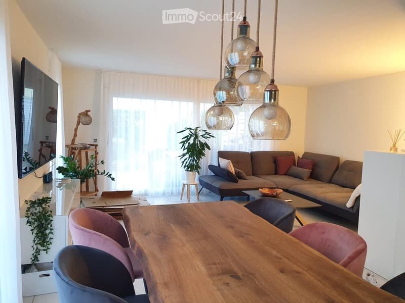 Modern living and dining area with a grey sectional sofa, wooden coffee table, large wooden dining table with pink chairs, pendant lights, and a TV screen on the wall.