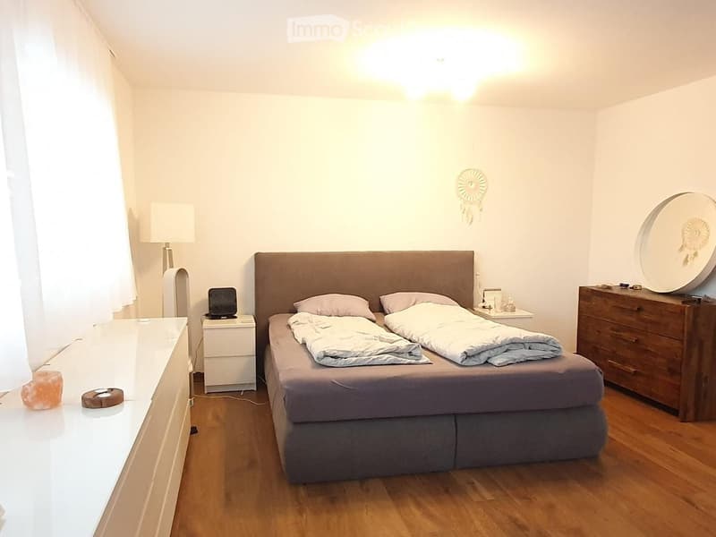 A cozy bedroom with a large bed, nightstand, lamp, and a wooden dresser. The room features wooden flooring and white walls.
