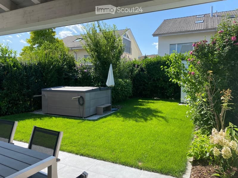Well-maintained garden with a small hot tub, lush green lawn, and surrounding trees and shrubs. A patio area with a table and chairs is visible in the foreground.
