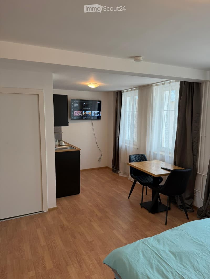 A modern, compact studio apartment with a bed, a small kitchen area, a dining table with chairs, and a mounted television. The room features wooden flooring and large windows with curtains.