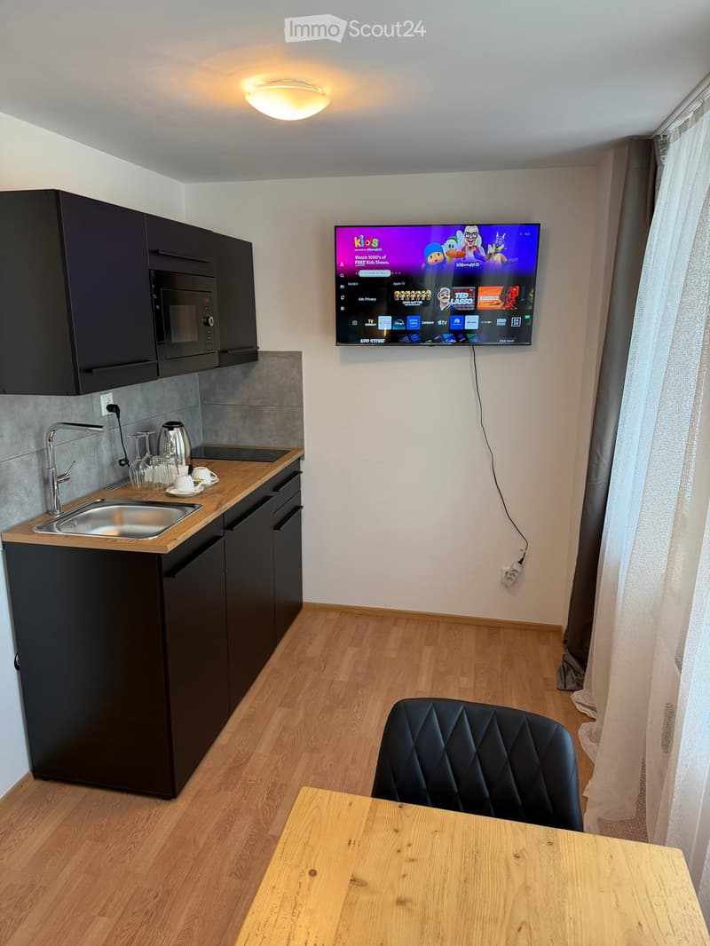 A modern, compact living space with a kitchen area and a living area. Features include a wooden table, a black quilted chair, a microwave, a sink with a faucet, and a mounted TV screen displaying a streaming service interface.