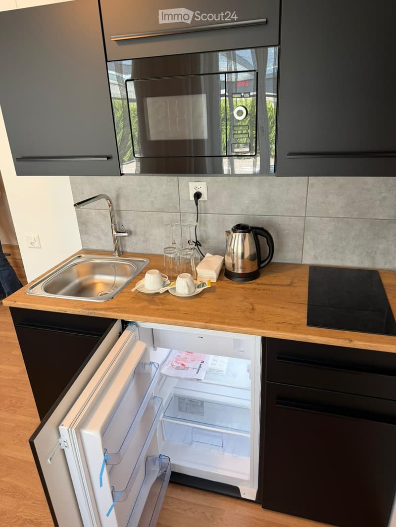 Modern kitchen with wooden countertops, stainless steel sink, microwave, and open refrigerator. Includes cups, kettle, and other small appliances.