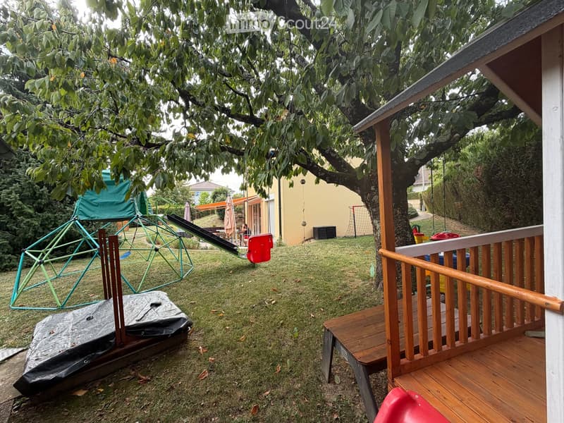 A well-maintained garden with a wooden deck, playground equipment, and a covered seating area under a large tree.