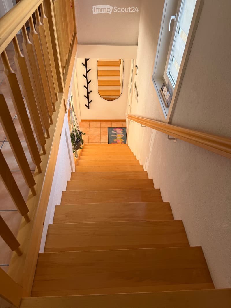 Wooden staircase with a wooden handrail, beige walls, and a small window on the right side. A decorative branch and a small potted plant are visible on the landing.