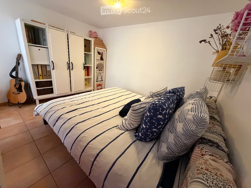 Cozy bedroom with a bed, striped duvet, pillows, a guitar leaning against a white wardrobe, and a small bookshelf with decorative items.