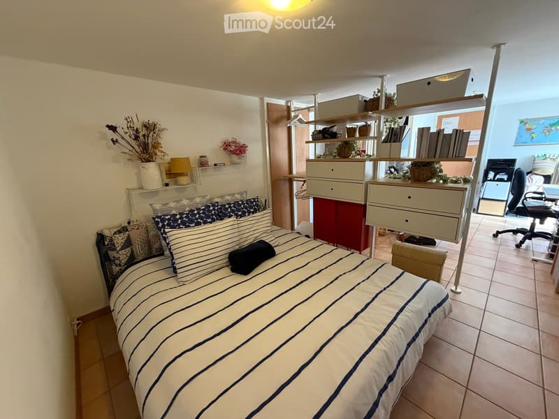 Cozy bedroom with a single bed, striped bedding, nightstand, shelves with decorative items, and an adjacent workspace area with a desk and office chair.