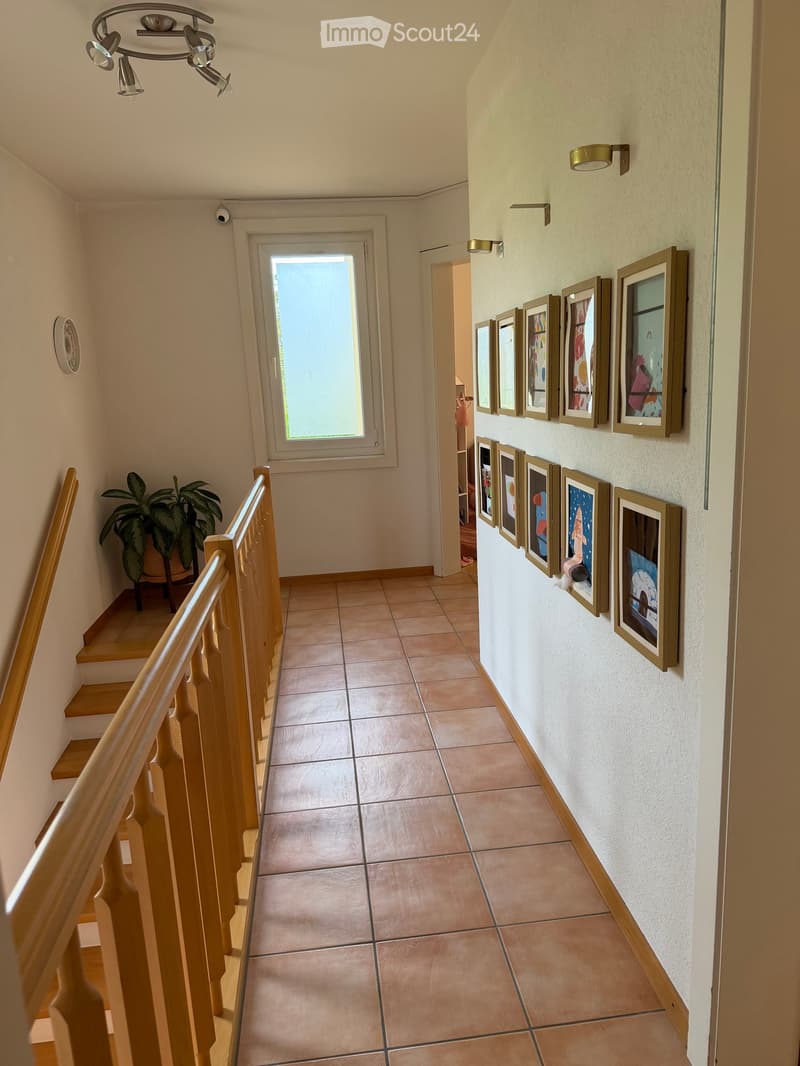 Hallway with tiled floor, wooden staircase railing, framed artwork on walls, and a potted plant near the stairs.