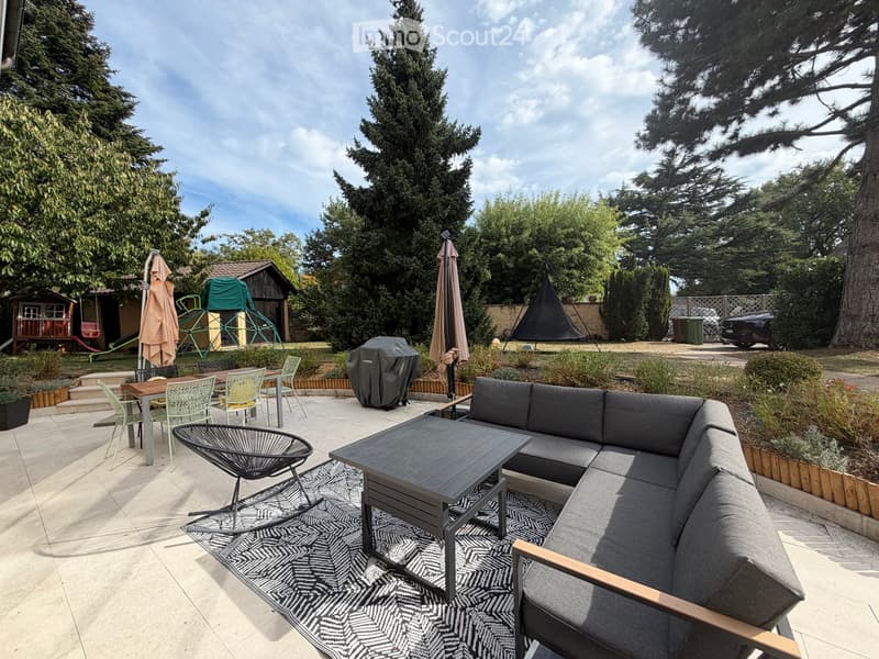 Outdoor patio with a grey sectional sofa, coffee table, dining table with chairs, umbrella, covered grill, and a small playground in the background.
