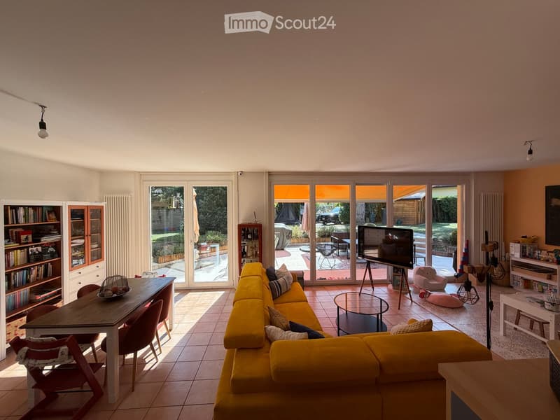 Bright living room with large windows and sliding glass doors leading to a patio. Features a yellow sectional sofa, wooden dining table with chairs, bookshelf, and a TV on a stand.