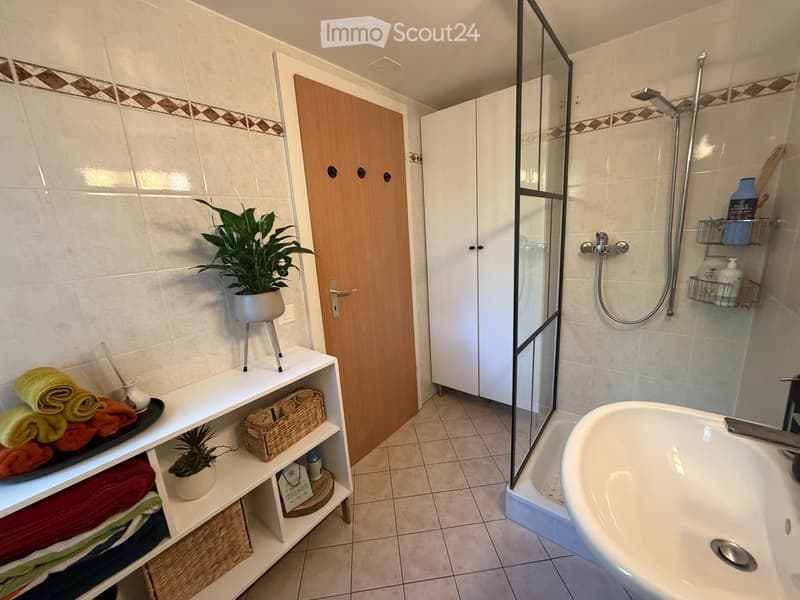 Modern bathroom with a bathtub, glass-enclosed shower, white tiled walls, and a wooden door. Shelves hold towels, plants, and decorative items.