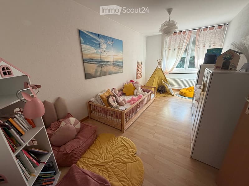 A well-lit children's bedroom with a wooden bed, colorful pillows, a bookshelf, a round yellow rug, a small sofa, and a toy tent in the corner.