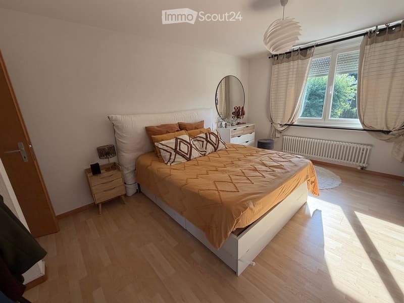 Bright bedroom with wooden flooring, large bed with brown bedding, nightstand with lamp, mirror, and window with curtains.