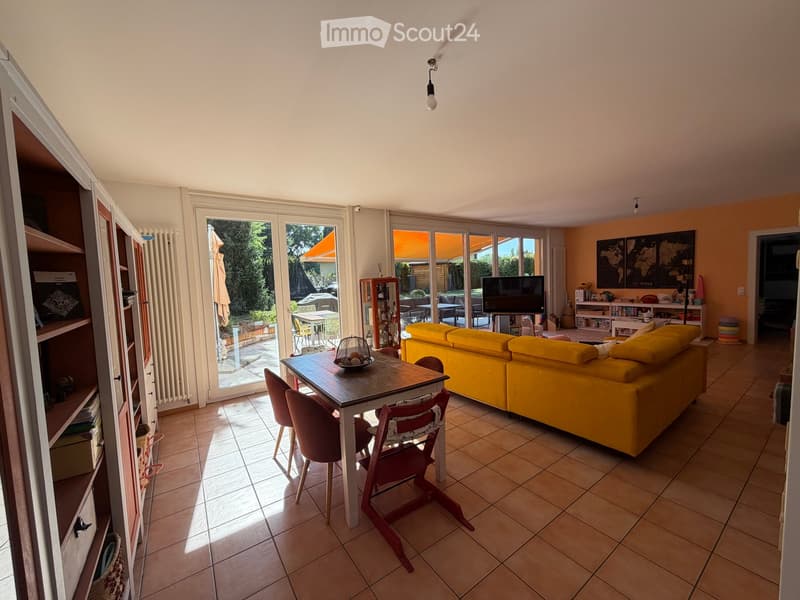 Bright living room with large glass doors leading to a patio, yellow sofa, wooden dining table with chairs, bookshelf, and television.