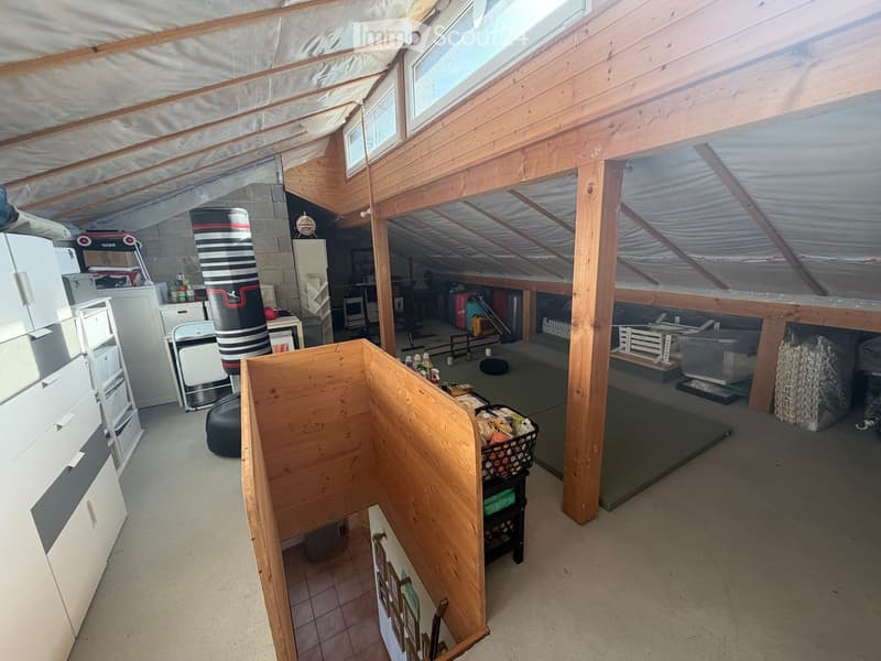 The attic space is furnished with a refrigerator, microwave, punching bag, chairs, a table, a basket, a stool, and shelves. The room features wooden beams and skylights, providing ample natural light.