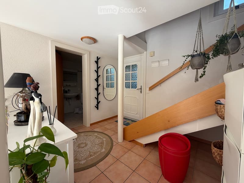 Hallway with a staircase leading to an upper level, featuring a white table with a lamp, a decorative figurine, and hanging plants. A doorway leads to a bathroom with a sink and toilet visible.