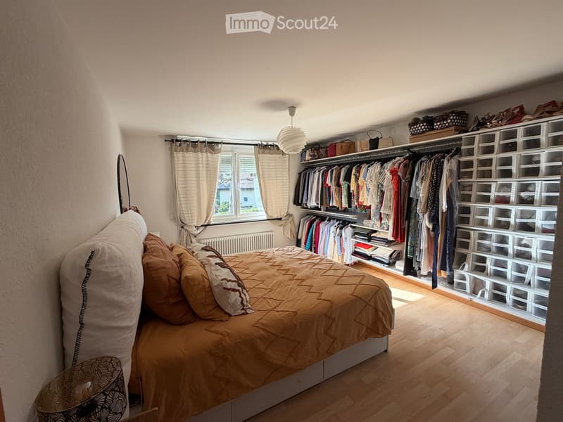 Cozy bedroom with a large bed, pillows, and a wardrobe with hanging clothes and storage boxes. The room features wooden flooring, a window with curtains, and a modern lamp.