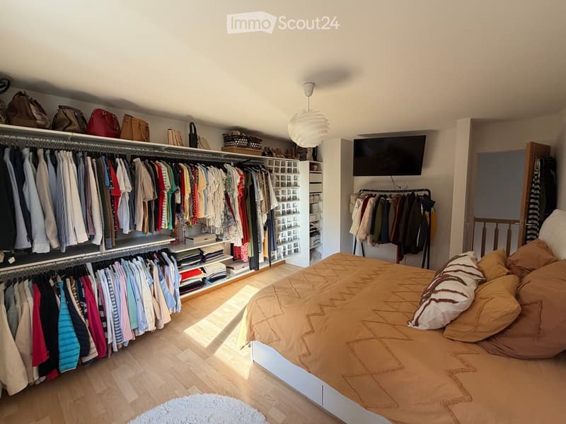 Well-lit bedroom with a large bed, a built-in wardrobe, and a clothing rack. The room features a pendant light, a screen mounted on the wall, and several handbags and baskets on the wardrobe shelves.