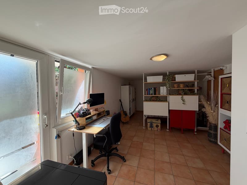 A well-lit room with a desk setup, a guitar, shelves, and storage boxes. The room features tiled flooring and large windows with blinds.