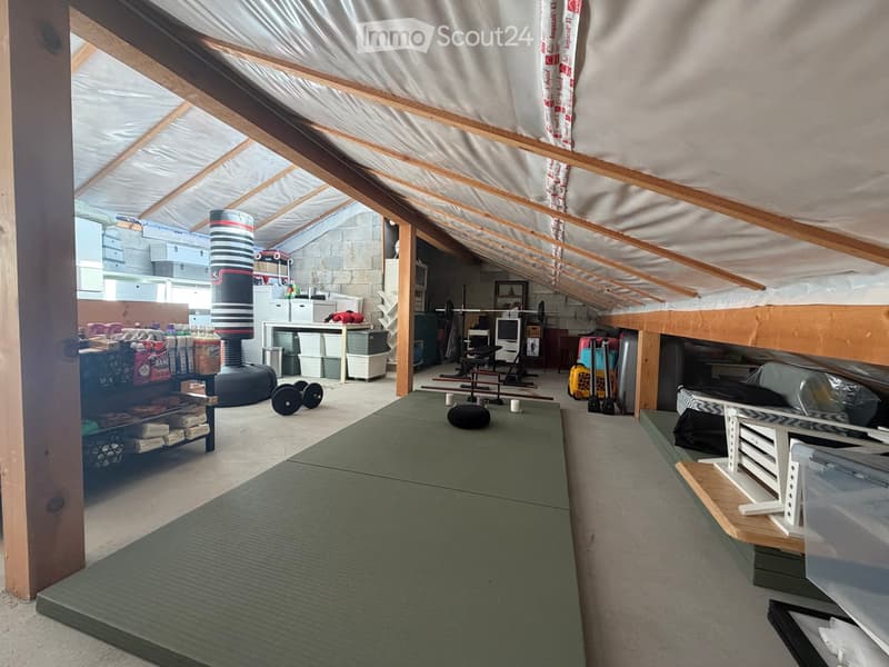 An attic space with exposed wooden beams, featuring a lofted bed area, a workout area with weights and a mat, and storage bins and shelves.