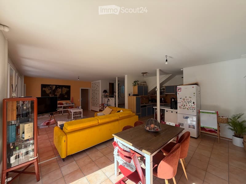 Bright living and dining area with yellow sofa, wooden dining table, colorful chairs, kitchen area with cabinets and refrigerator, and decorative screen easel.