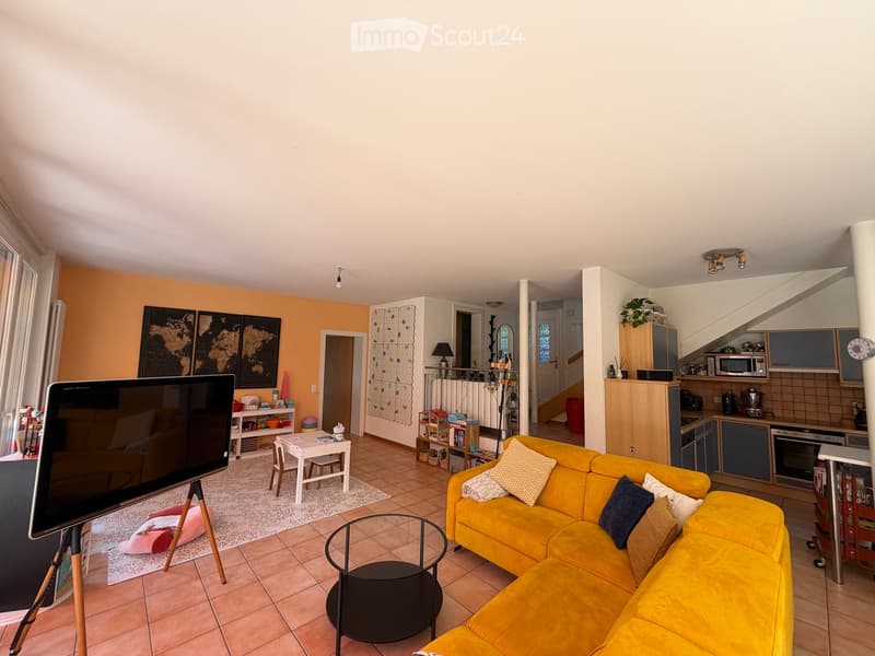 Bright living room with a yellow sofa, coffee table, small dining table, and television. The space features a kitchen area with appliances and a staircase leading to another level.