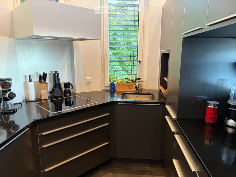 Modern kitchen with dark countertops, stainless steel appliances, and sleek cabinetry. Features include a coffee maker, blender, knife block, and a sink with a window view.
