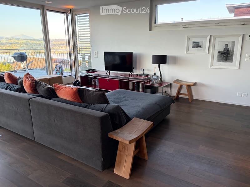 Modern living room with large windows offering a scenic view, featuring a grey sectional sofa, wooden stools, a coffee table, a floor lamp, and a television on a red and silver stand.
