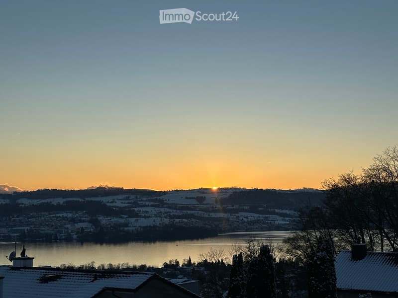 Panoramic view of a snow-covered landscape at sunset, featuring a lake, distant mountains, and rooftops in the foreground.