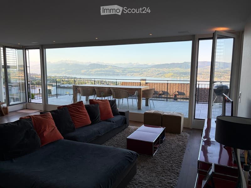 Modern living room with large windows offering a scenic mountain view, featuring a dark grey sectional sofa with colorful pillows, a sleek coffee table, and a small outdoor dining area with chairs and a table.