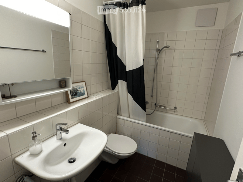 Modern bathroom with white tiles, a sink, toilet, bathtub, shower, and a mirror cabinet. Features a black and white shower curtain and a framed picture on the shelf.