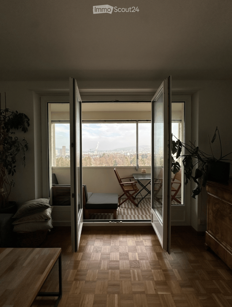 Modern living room with wooden flooring, large windows offering a city view, and a small seating area with a sofa, table, and chairs.