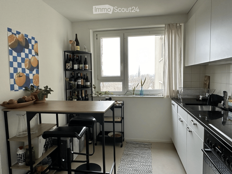 Modern kitchen with white cabinets, a wooden table, two black stools, a sink with a faucet, an oven, a bottle rack, a bowl of fruit, a vase with a plant, and a painting on the wall.