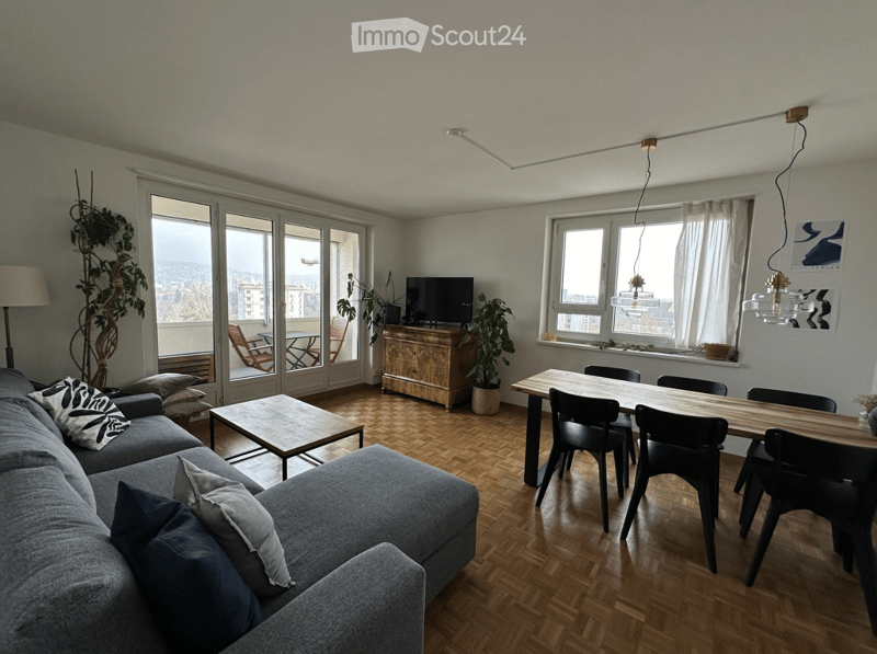 Modern living and dining area with wooden flooring, large windows, a grey sectional sofa, coffee table, dining table with chairs, TV, and potted plants.