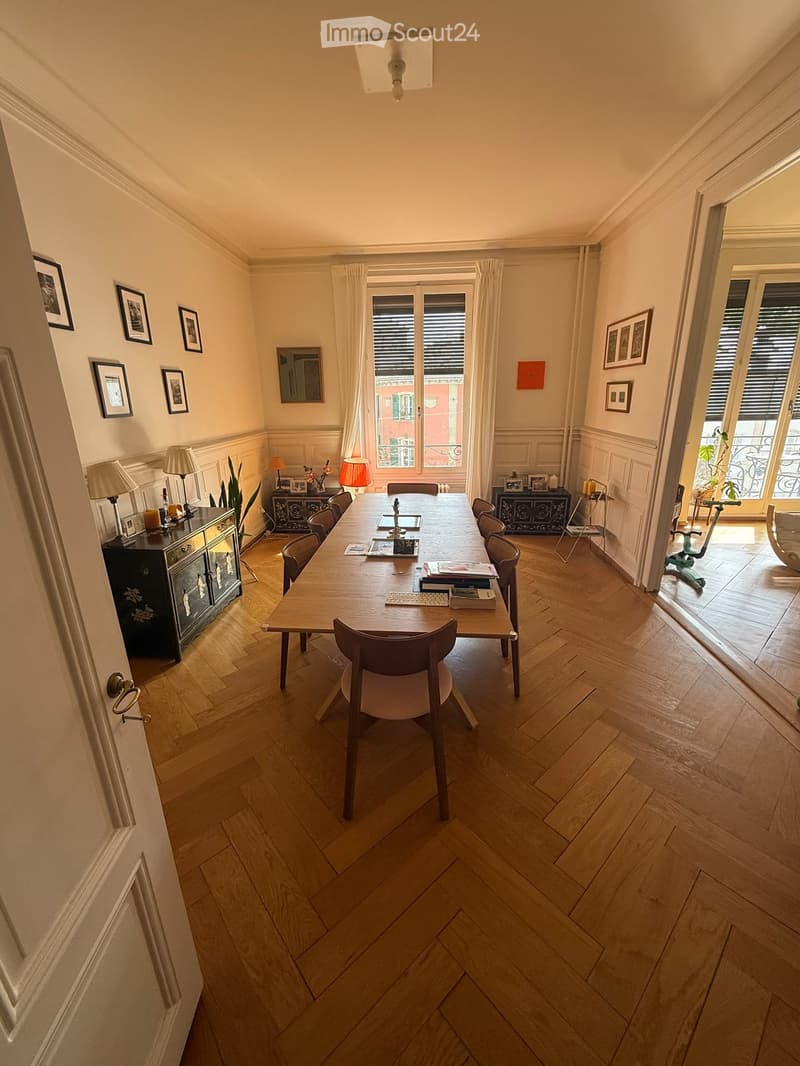 Spacious room with wooden herringbone flooring, large table surrounded by chairs, cabinets with lamps, framed pictures on walls, and a view of a balcony.