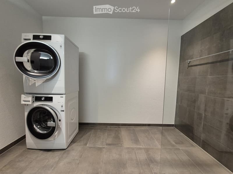 A small, modern laundry room with a stacked washer and dryer unit, tiled floor, and a glass shower enclosure.