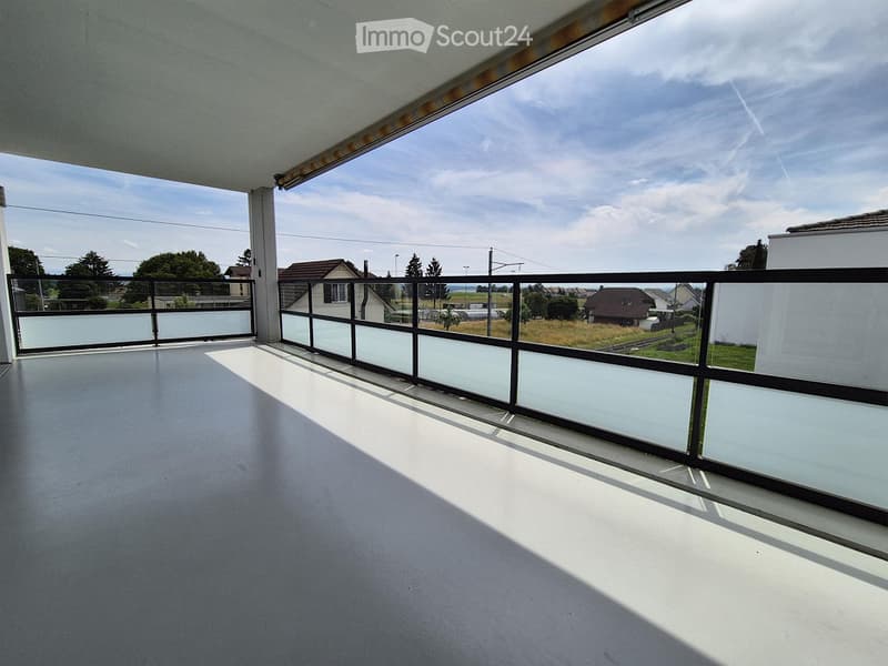 Empty balcony with modern glass railing, concrete floor, and scenic view of surrounding houses and fields.