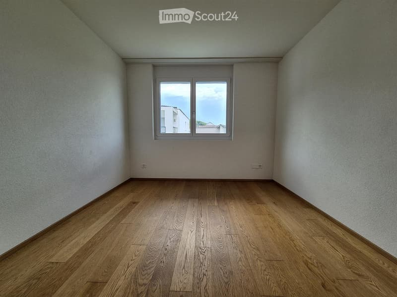 Empty room with wooden flooring and white walls, featuring a large window with a view of neighboring buildings.
