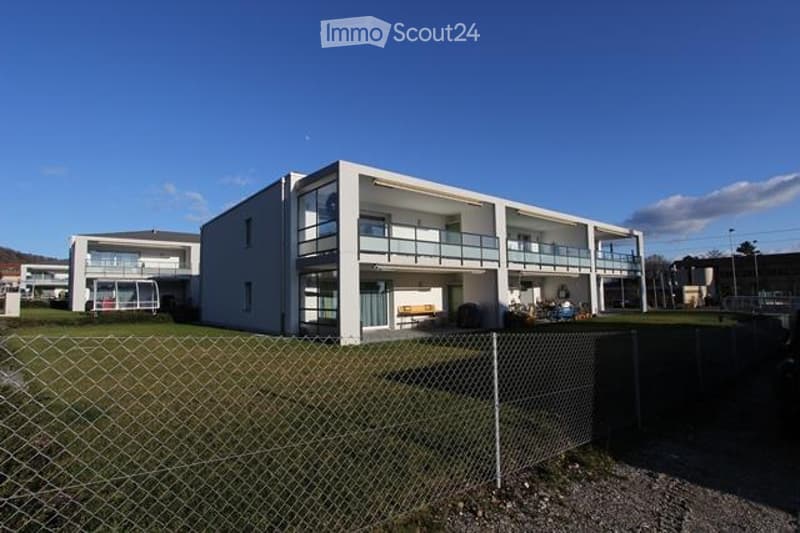 Modern two-story building with white exterior, multiple windows, and balconies. The building is surrounded by a fenced area with grass and gravel.
