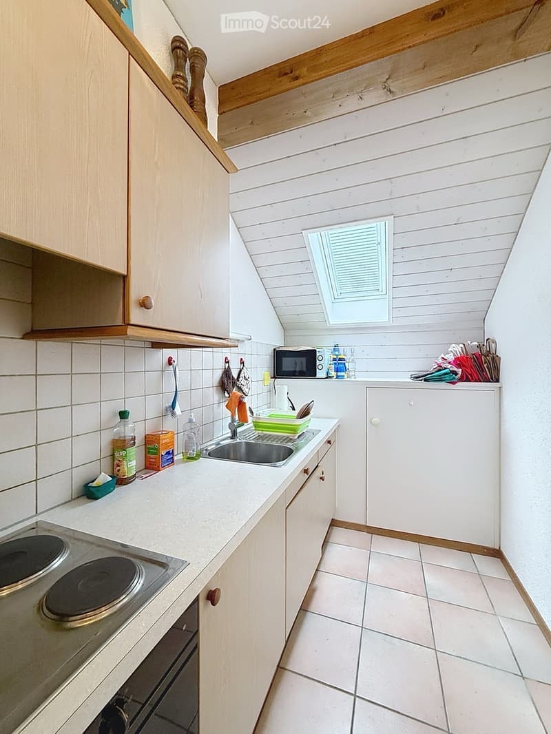 The kitchen features white tiled walls and a white tiled floor. It includes a sink with a faucet, a microwave, an oven, and light wooden cabinets. Various items such as bottles, cups, a bowl, and an oven mitt are placed on the countertops.