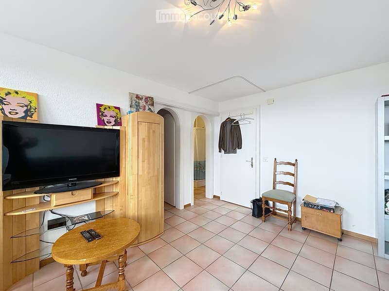 A furnished room with a television on a wooden stand, a wooden cabinet, a small round wooden table with remote controls, a wooden chair, and several artworks on the walls. The room has tiled flooring and white walls.