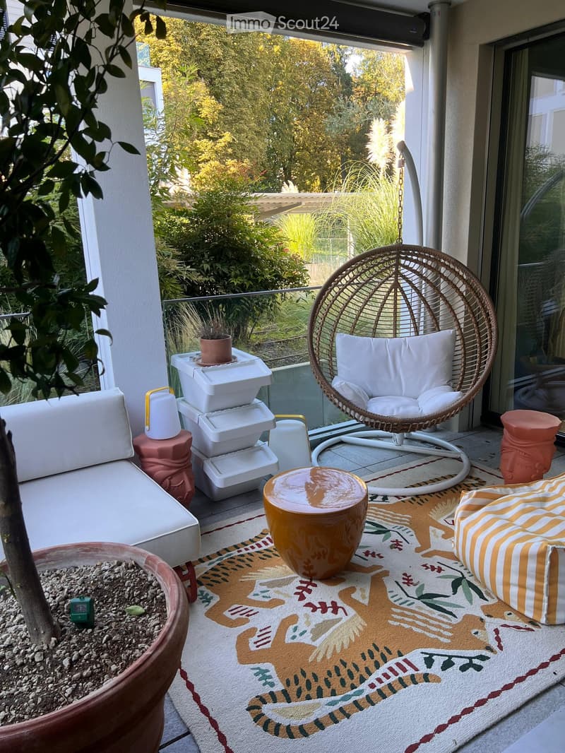 Balcony with a white sofa, wicker hanging chair, small table, colorful rug, and potted plants.