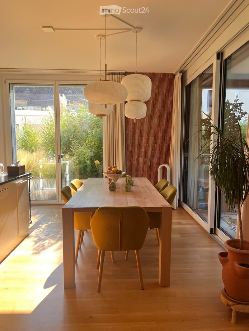Bright dining area with a long wooden table, six green chairs, a fruit bowl, and a large potted plant near the window. The room features modern pendant lights and large glass doors leading to a garden.