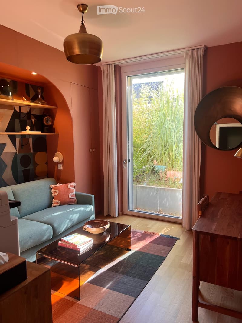 Modern living room with a blue sofa, coffee table, books, and a view of greenery through large windows.