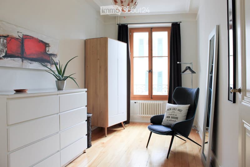 A modern, well-lit room with light wooden flooring, a large window with dark curtains, a sleek black chair with a decorative pillow, a tall white cabinet, a potted plant on top of a white dresser, and a large abstract painting on the wall.